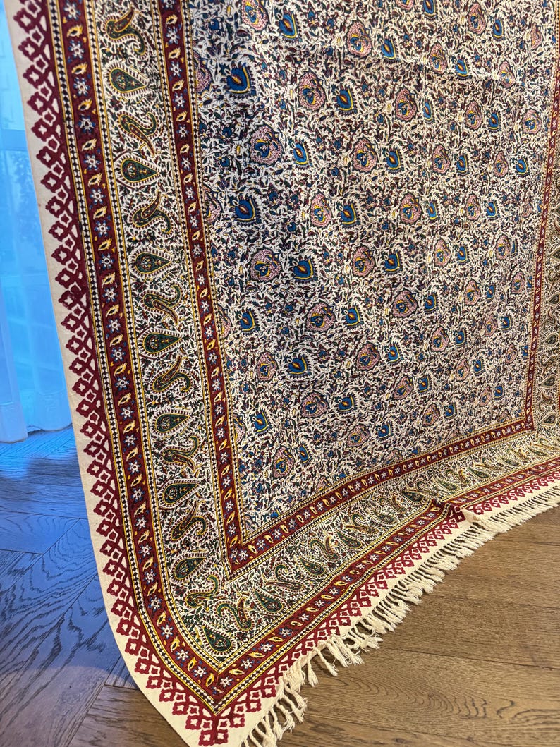 May include: A rectangular, patterned rug with a cream-colored background. The rug features a repeating floral design in shades of blue, pink, and green. The border is a deep red with geometric and paisley patterns, and the rug has a fringed edge.