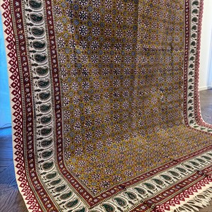 May include: A patterned textile with a mustard yellow background and a repeating floral design. The border features a paisley pattern in green, red, and white, with a fringed edge. The textile is likely a rug or a wall hanging.