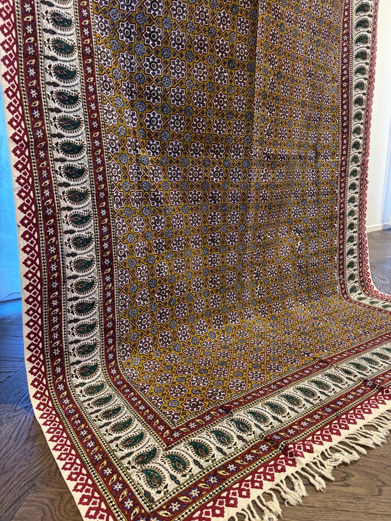May include: A patterned textile with a mustard yellow background and a repeating floral design. The border features a paisley pattern in green, red, and white, with a fringed edge. The textile is likely a rug or a wall hanging.
