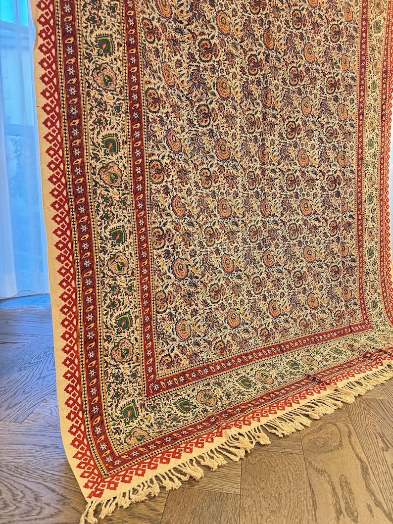 May include: A cream-colored textile with a detailed floral pattern in shades of red, orange, green, and blue. The border features a red geometric design and fringe. This decorative textile is suitable for a variety of uses.