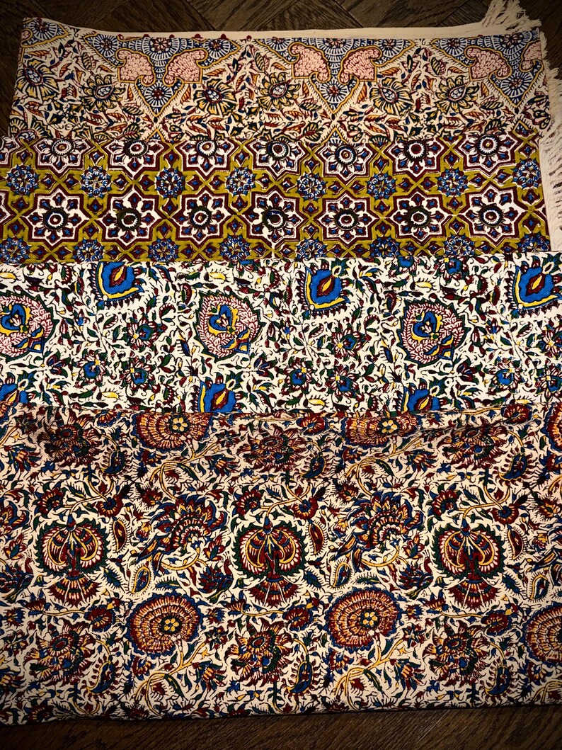 May include: A folded textile with intricate floral and geometric patterns in shades of red, blue, yellow, and white. The design includes repeating floral motifs and star-like shapes. The textile has a fringed edge.
