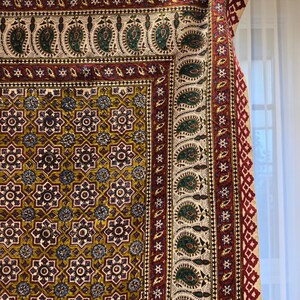 May include: A detailed shot of a patterned textile with a complex design. The fabric features a repeating floral and geometric pattern in shades of gold, burgundy, green, and blue. The edges are trimmed with a fringe. The textile is likely a rug or tapestry.