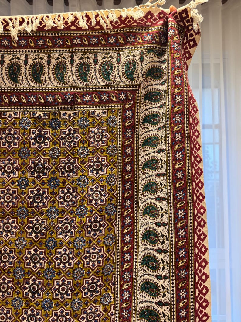 May include: A detailed shot of a patterned textile with a complex design. The fabric features a repeating floral and geometric pattern in shades of gold, burgundy, green, and blue. The edges are trimmed with a fringe. The textile is likely a rug or tapestry.