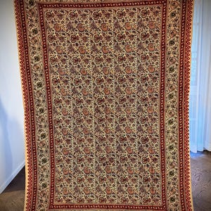May include: A large, rectangular beige textile with a repeating floral pattern in shades of red, brown, and green. The textile has a red border with a geometric design and fringe along the bottom edge. The textile is hanging.