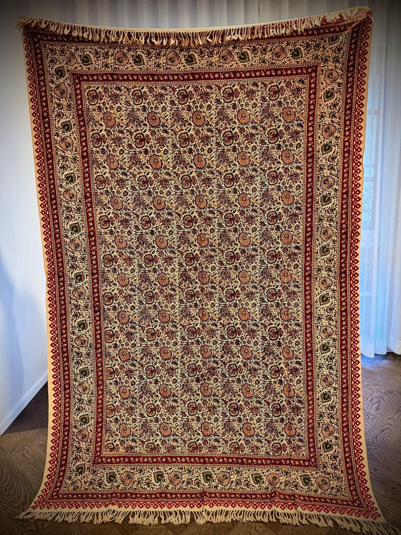 May include: A large, rectangular beige textile with a repeating floral pattern in shades of red, brown, and green. The textile has a red border with a geometric design and fringe along the bottom edge. The textile is hanging.
