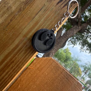 May include: A black circular keychain featuring a raised musical note symbol on the front surface. The keychain is attached to a silver-colored metal chain and a circular split ring, showcasing a textured finish from the 3D printing process.