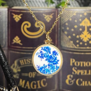 Könnte beinhalten: Eine goldene Kettenkette mit einem runden Anhänger, der mit einem blauen und weißen Blumenmuster verziert ist. Der Anhänger ist von einem goldenen Rand umgeben. Die Kette hängt an einem Ast, auf dem zwei schwarze Vögel sitzen. Der Hintergrund ist ein Bücherregal mit Büchern mit den Titeln "Geschichte der Magie" und "Tränke, Zauber und Reize".