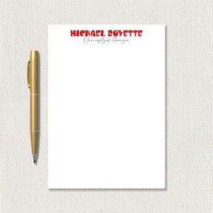 May include: A white notepad with the name "MICHAEL ROYETTE" in red and "University of Georgia" in black script. A gold pen is positioned to the left of the notepad.