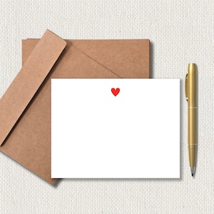May include: A white stationery card with a small red heart, a brown envelope, and a gold pen. The card and envelope are on a textured, off-white surface. The pen is positioned vertically to the right of the card.