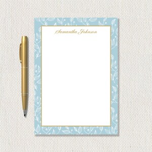 May include: A light blue notepad with a white floral design border and a gold border. The notepad has the name "Samantha Johnson" printed in gold script. A gold pen is to the left of the notepad.