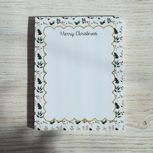May include: A white notepad with a gold border and a holly leaf pattern. The notepad has the words "Merry Christmas" printed on the top.