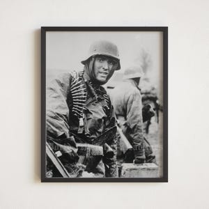 May include: Black and white photograph of a soldier in a military uniform and helmet. The soldier is holding a rifle and looking directly at the camera. The soldier is standing in front of another soldier who is out of focus.