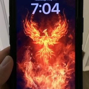 Könnte beinhalten: Ein Smartphone mit schwarzem Gehäuse, das ein Hintergrundbild mit einem feuerspeienden Phönix zeigt. Die Uhrzeit ist 7:04 Uhr und das Datum ist Dienstag, der 14. Mai.