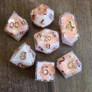 May include: A set of seven polyhedral dice featuring a translucent white and peach-colored resin base with embedded iridescent flakes. Each die has engraved numbers filled with a metallic copper-toned ink, displaying various geometric shapes.