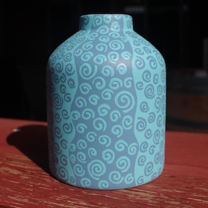 May include: A cylindrical vase featuring a matte finish with a light blue base colour and a repeating pattern of darker blue hand-painted swirls. The vase has a narrow neck and a wide, rounded body, displaying a consistent colour palette throughout.