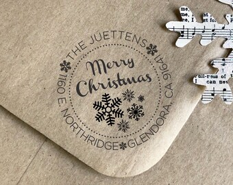 May include: A kraft paper envelope with a circular "Merry Christmas" stamp featuring snowflakes. The stamp includes the address "1160 E. Northridge Glendora, CA 91741". A snowflake cut from sheet music is placed on the envelope.