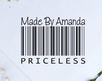 May include: A white rectangular card featuring the black text Made By Amanda above a vertical barcode graphic, with the word PRICELESS printed in black capital letters below the barcode, set against a background with white pearls and green floral stems.