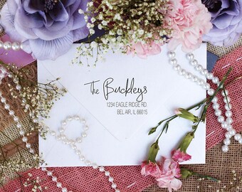 May include: A white envelope with a return address label that reads "The Buckleys, 1234 Eagle Ridge Rd., Bel Air, IL 66015". The envelope is surrounded by pink and purple flowers, white pearls, and burlap fabric.