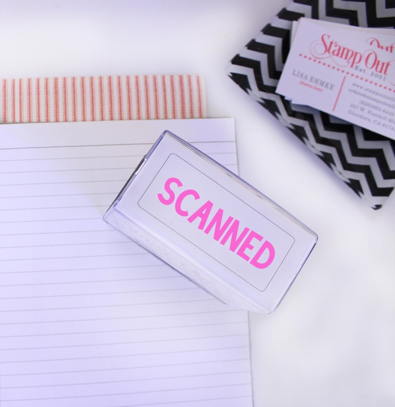 May include: A clear plastic stamp with the word "SCANNER" in pink letters. The stamp is on a white surface with a lined notebook and a business card.