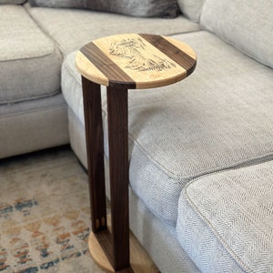 May include: A round wooden side table with a walnut and maple wood top. The table has a unique design with a wood-burned image of a mountain range. The table is next to a grey sofa.