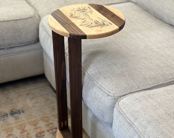 May include: A round wooden side table with a walnut and maple wood top. The table has a unique design with a wood-burned image of a mountain range. The table is next to a grey sofa.