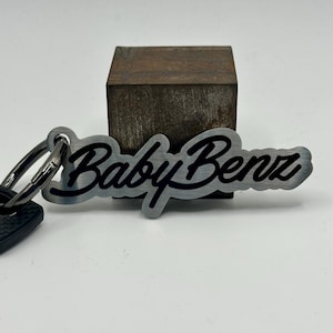 Puede incluir: Un llavero de metal plateado con las palabras "Baby Benz" en una fuente de escritura negra. El llavero está sujeto a un llavero plateado. El fondo es neutro.