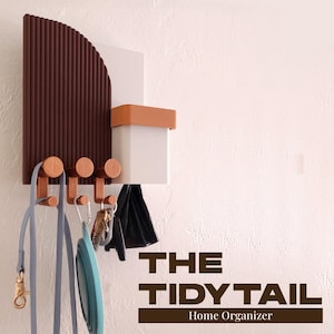 May include: A wall-mounted home organiser with a brown, ridged section, a white square, and a small tan-coloured box. It features wooden hooks holding a lead, keys, and a collapsible bowl. The text "THE TIDY TAIL Home Organizer" is displayed.