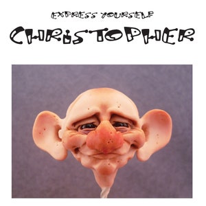 Puede incluir: Un primer plano de una cara de arcilla esculpida con una expresión feliz. La cara es de color marrón claro con una nariz rosa y ojos oscuros. La cara está unida a una varilla delgada y blanca.