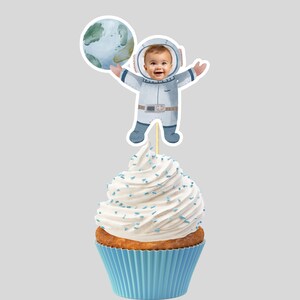 Op de afbeelding: Een blauwe en witte astronaut cupcake topper met een foto-uitsparing voor een gezicht. De astronaut houdt een blauwe en groene planeet vast. De topper zit bovenop een witte cupcake met blauwe sprinkles.