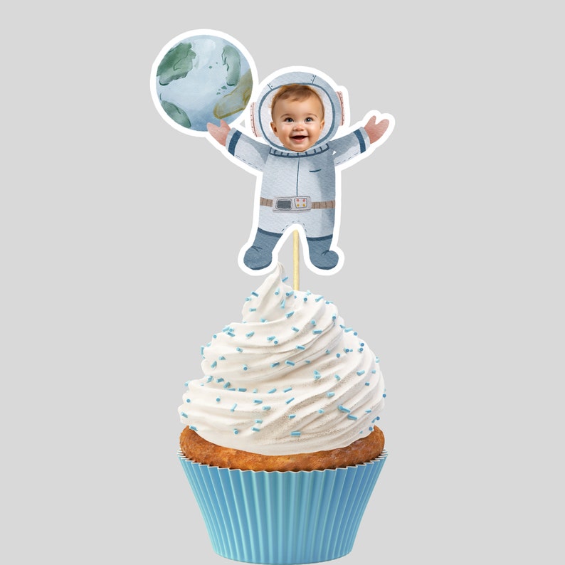 Op de afbeelding: Een blauwe en witte astronaut cupcake topper met een foto-uitsparing voor een gezicht. De astronaut houdt een blauwe en groene planeet vast. De topper zit bovenop een witte cupcake met blauwe sprinkles.