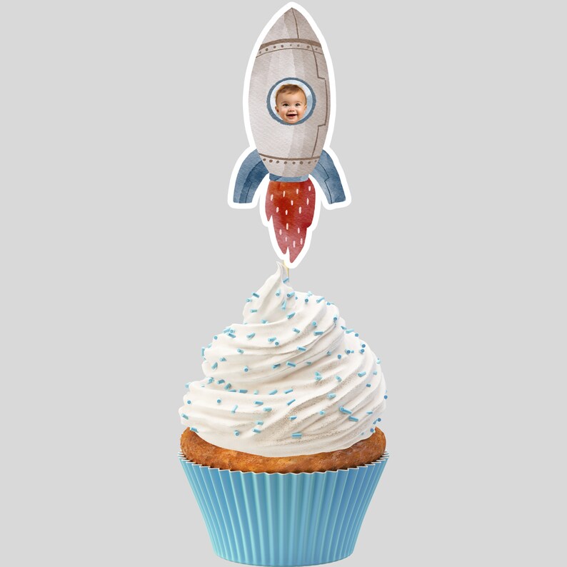 Op de afbeelding: Een witte cupcake met blauwe sprinkles, bedekt met een raket cupcake topper. De raket is grijs met een rode vlam en een blauw raam. Een kindergezicht is zichtbaar in het raam.