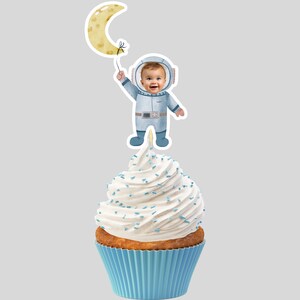 Op de afbeelding: Een blauwe en witte cupcake met witte frosting en blauwe sprinkles, bedekt met een cupcake topper van een cartoon astronaut die een gele halve maan ballon vasthoudt. De astronaut heeft een ruimtehelm met een uitsparing voor een gezicht.
