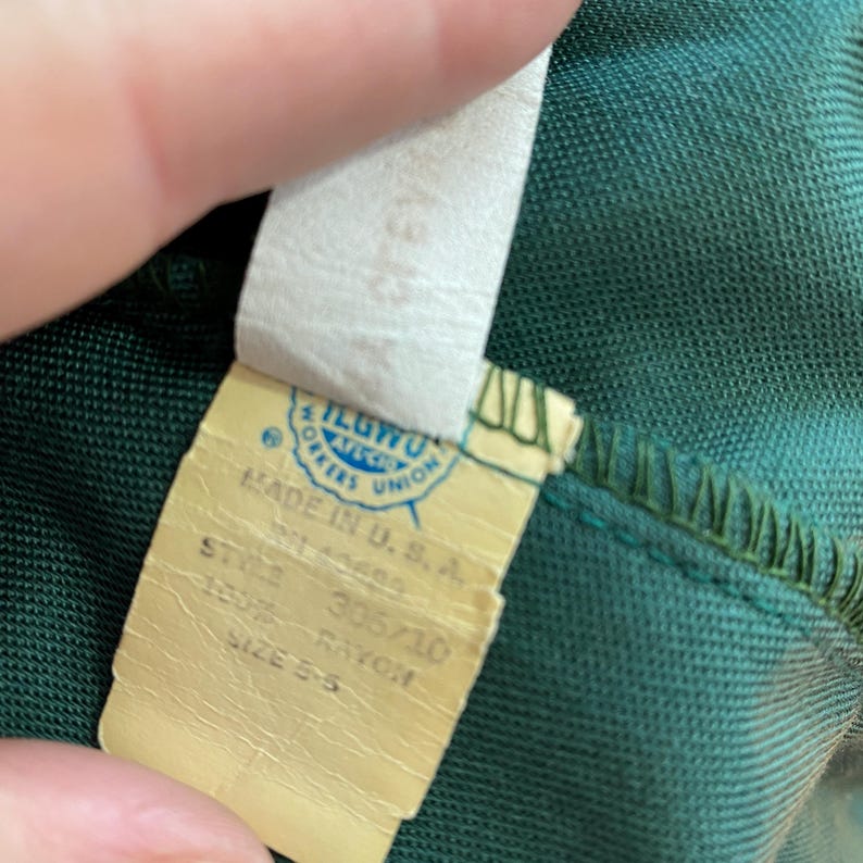 Puede incluir: Una etiqueta de tela verde con texto blanco que dice "Made in U.S.A.", "Style 305/10", "100% Rayon", y "Size 55". La etiqueta tambi&eacute;n tiene una etiqueta circular azul y blanca que dice "ILGWU AFL-CIO Workers Union".