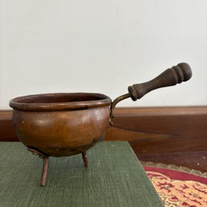 May include: A small, antique copper pot with three legs and a wooden handle. The pot has a rounded bowl shape and a dark brown patina. The handle is attached with a brass fitting.