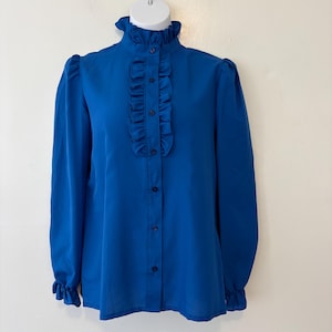 May include: A vibrant blue blouse with a high ruffled collar and long sleeves. The front features a vertical ruffle detail and is fastened with small, dark blue buttons. The sleeves end in ruffled cuffs, creating a classic, elegant look.