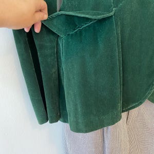 Puede incluir: Una chaqueta de terciopelo verde oscuro con un detalle de volantes en la espalda. La chaqueta est&aacute; desabrochada y la tela tiene una textura suave y aterciopelada.