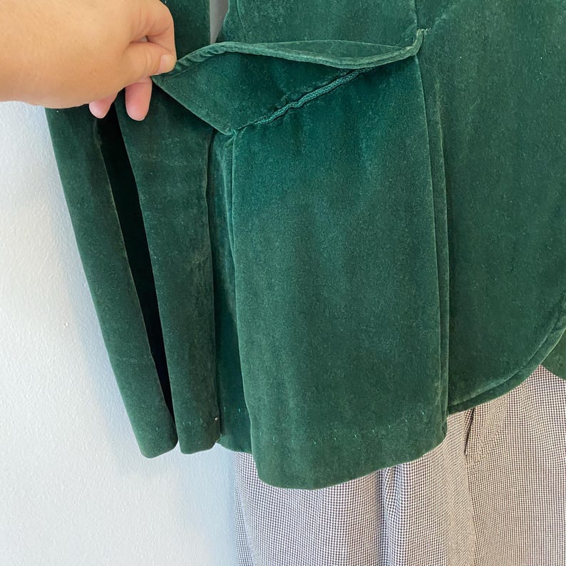 Puede incluir: Una chaqueta de terciopelo verde oscuro con un detalle de volantes en la espalda. La chaqueta est&aacute; desabrochada y la tela tiene una textura suave y aterciopelada.