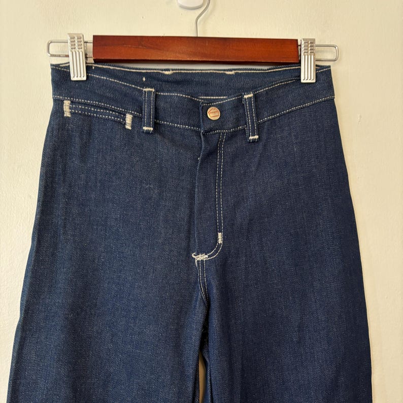 May include: Dark blue denim pants with a button closure and white stitching. The pants are hanging on a wooden hanger. The pants have a high waist and a straight leg.