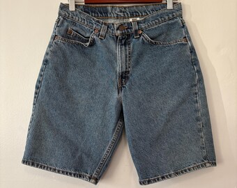 May include: Blue denim shorts with a classic five-pocket design. The shorts are hanging on a wooden hanger against a white wall. The shorts have a button and zipper closure, and the denim has a faded appearance.