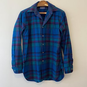 Pu&ograve; includere: Una camicia a quadri a maniche lunghe nei toni del blu, verde acqua e viola. La camicia ha un'abbottonatura frontale, un colletto e una tasca sul petto. La camicia &egrave; appesa a una gruccia di legno.