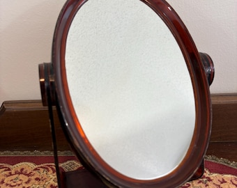 May include: A tabletop vanity mirror with an oval glass surface encased in a dark brown, translucent tortoiseshell-patterned frame. The mirror is mounted on a matching brown stand with two side pivots that allow the mirror to tilt and rotate.