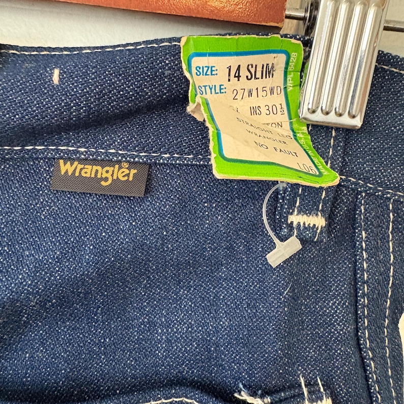 May include: Close-up of dark blue denim Wrangler jeans. The jeans have a tag that reads "SIZE: 14 SLIM" and "STYLE: 27W15WD". The tag also indicates an inseam of 30.5 inches. The Wrangler logo is visible on a small black tag.