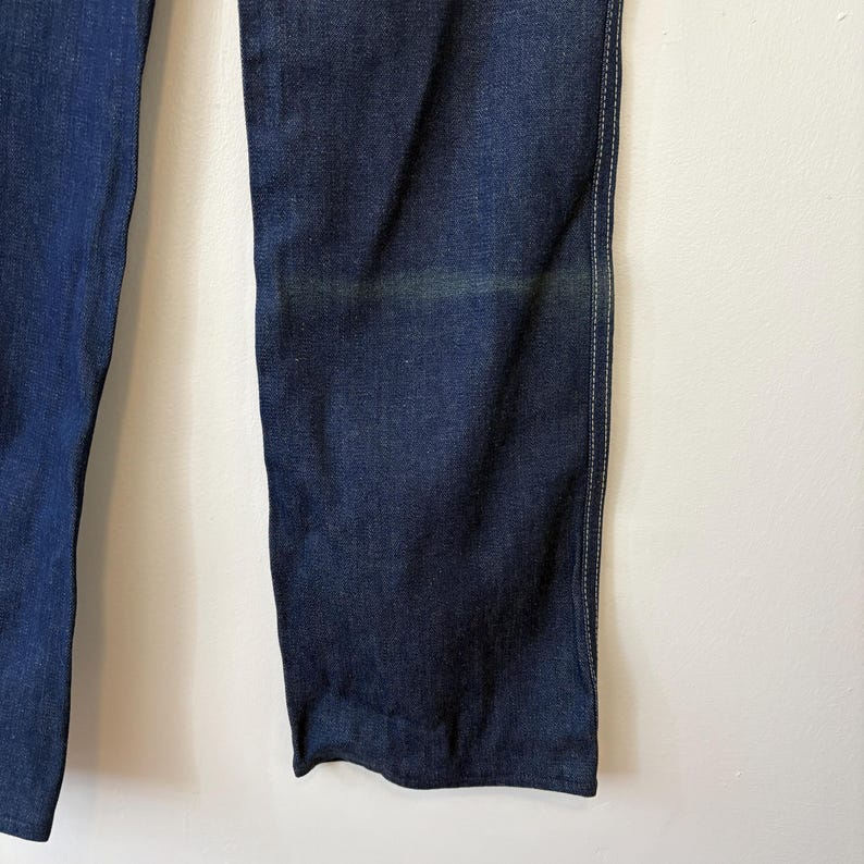 May include: Close-up of dark blue denim jeans. The jeans have a straight leg cut and are made of a sturdy denim material. The stitching is visible along the seams. The jeans are hanging against a white background.