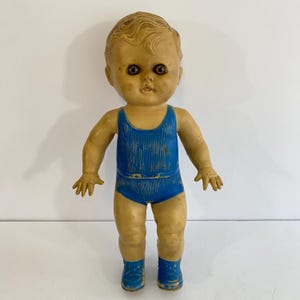 May include: Vintage plastic doll with a tan complexion, blue swimsuit, and matching blue shoes. The doll has molded hair and painted facial features, including dark eyes. The doll is standing upright, arms at its sides, against a white background.