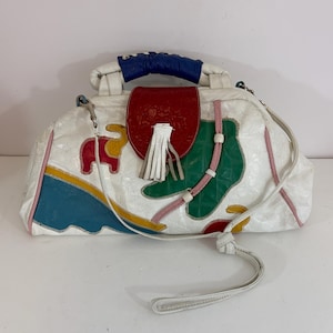 May include: White leather handbag with colorful patchwork design. Features a red flap with a white tassel, a blue handle, and a long white strap. The bag has a whimsical elephant and geometric shapes.