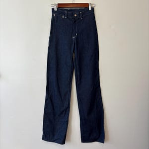 May include: Dark wash denim jeans hanging on a wooden hanger. The jeans have a classic five-pocket design, a button and zipper closure, and white stitching. The pants are straight-legged and appear to be a vintage style.