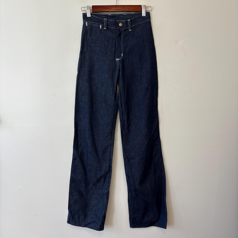 May include: Dark wash denim jeans hanging on a wooden hanger. The jeans have a classic five-pocket design, a button and zipper closure, and white stitching. The pants are straight-legged and appear to be a vintage style.