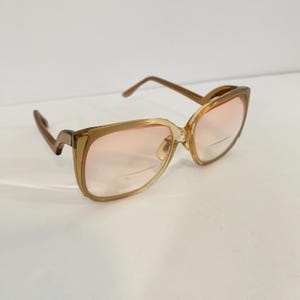 May include: Vintage square-framed eyeglasses with a light brown frame and pink-tinted lenses. The glasses have a classic design with a subtle cat-eye shape and a bridge. The lenses have a slight curve.