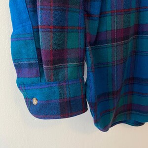 Pu&ograve; includere: Primo piano di una camicia di flanella a quadri blu, verde acqua e viola. La camicia presenta un polsino abbottonato e un dettaglio con un solo bottone. Il tessuto ha un aspetto strutturato, con un mix di colori e un classico motivo a quadri.