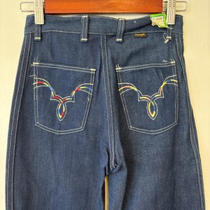 May include: Dark blue denim Wrangler jeans with white stitching and decorative back pockets. The pockets feature a colorful, embroidered design. The jeans are labeled "14 Slim".